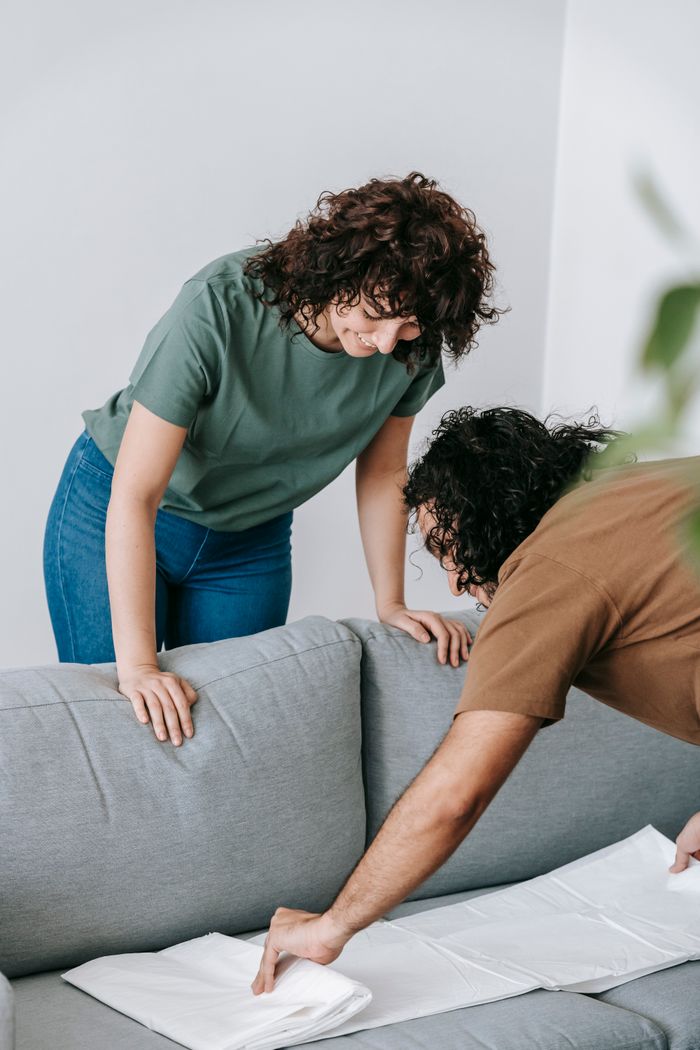 A couple getting the living room ready by draping the couch in a protective white cover.