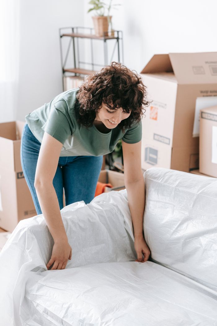 A woman is wrapping a sofa in plastic in a bright living room in preparation for moving.